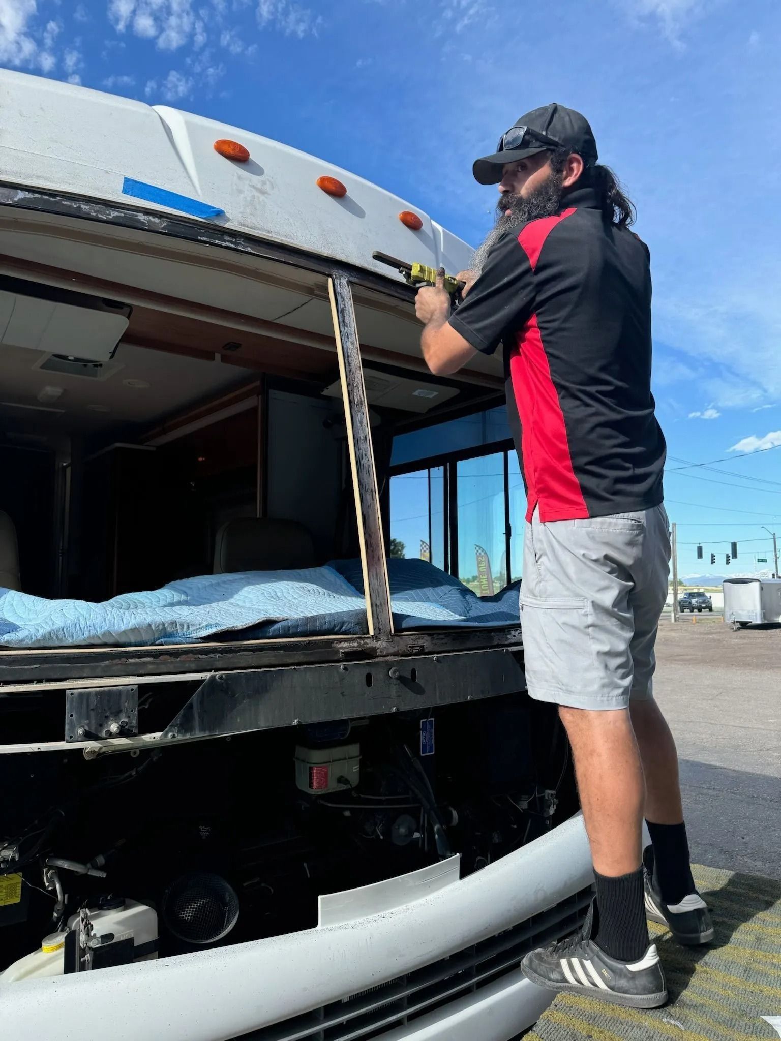 A person in a red and black shirt stands on the front bumper of a motorhome, using a power drill on the exterior.