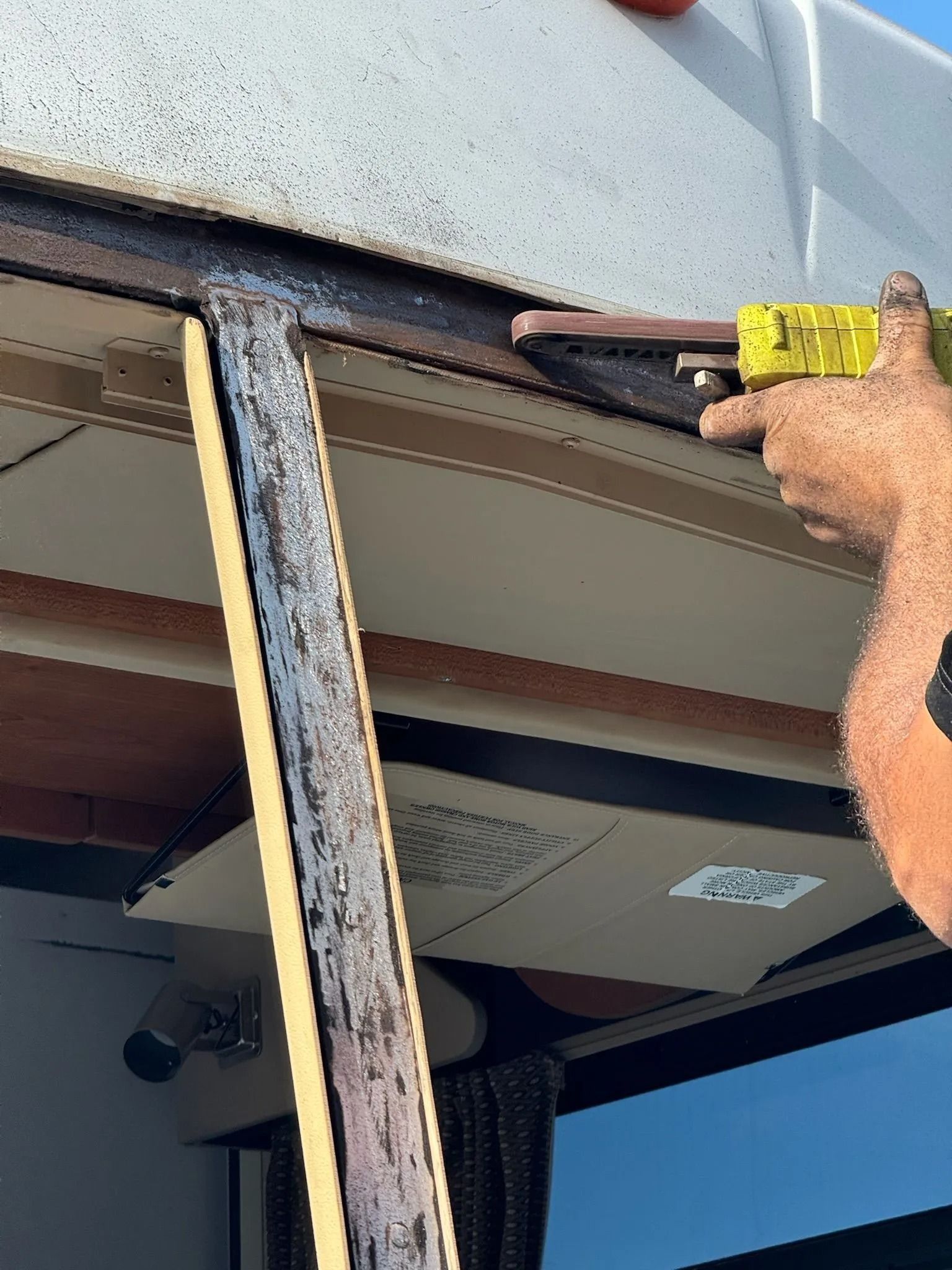 A person uses a power sander to smooth the edge of a structural metal beam on an RV or boat exterior.