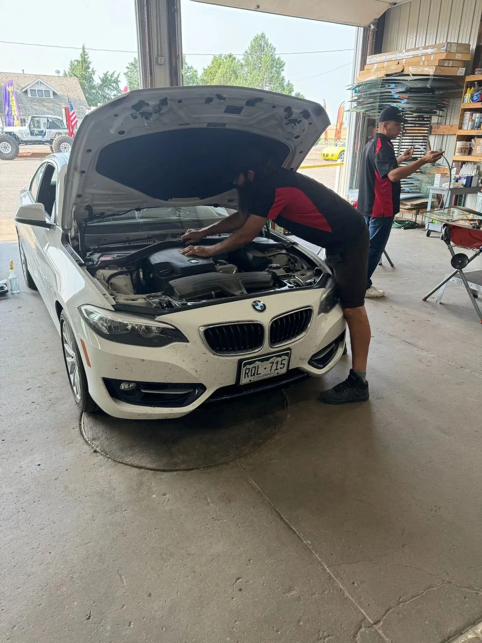 A mechanic works on the engine of a white BMW sedan inside a workshop, with another person standing in the background.
