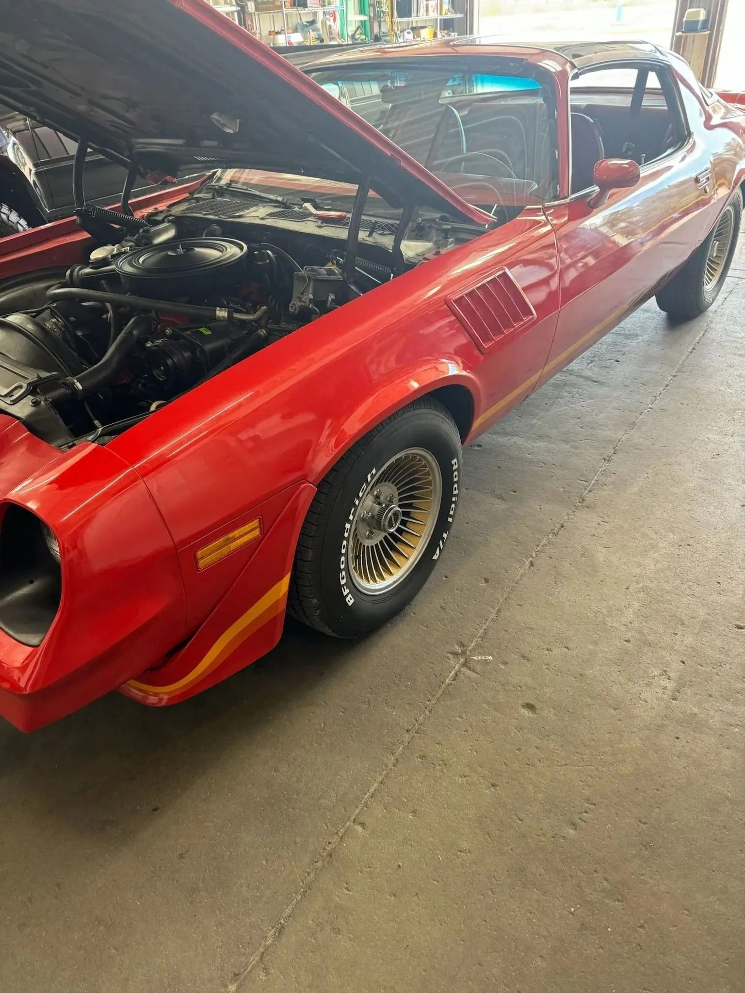 A red, vintage Pontiac Firebird with its hood open, parked on a concrete floor in a garage.