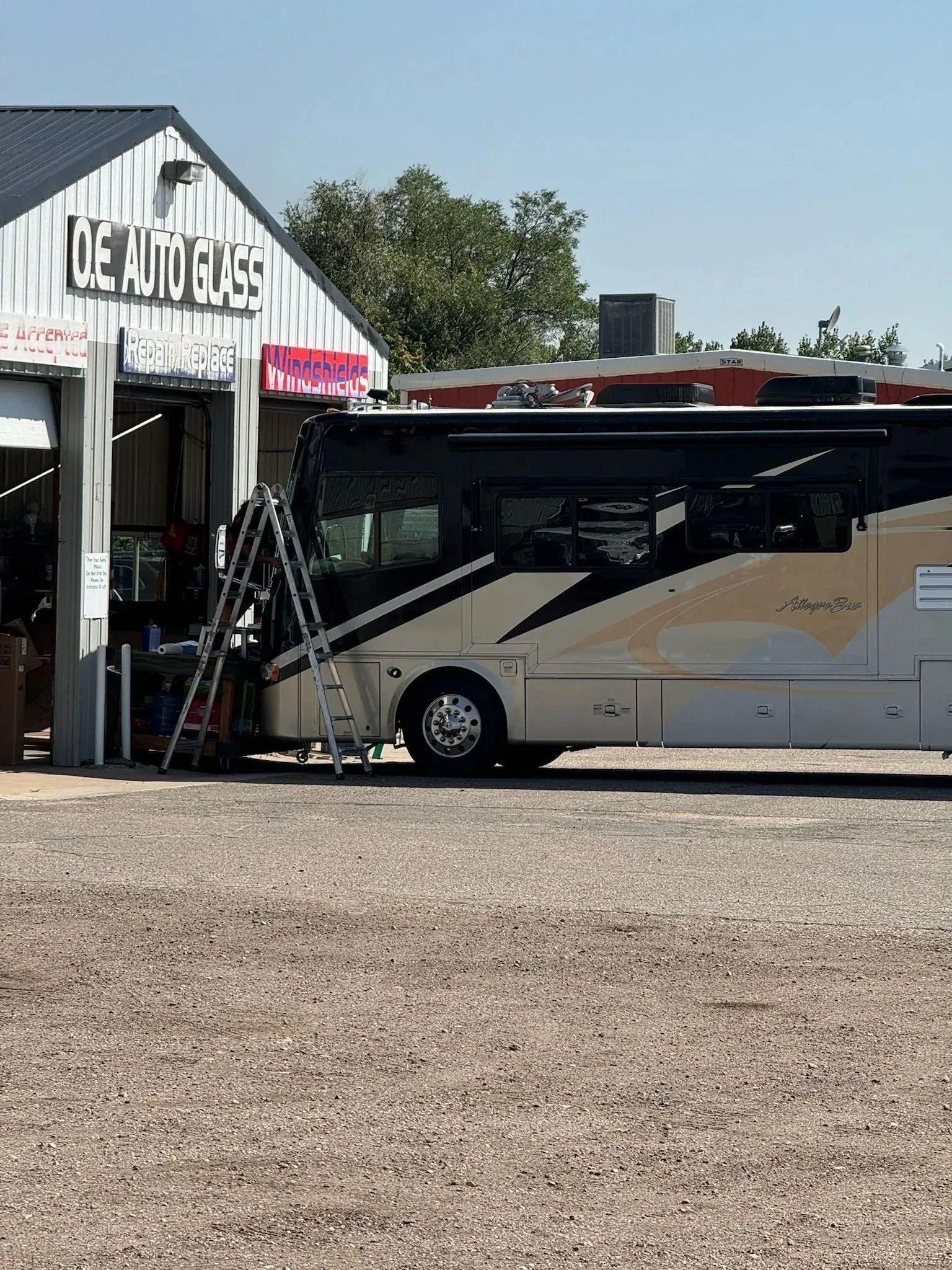 A large RV parked in front of an O.E. Auto Glass shop with a ladder leaning against its front windshield.