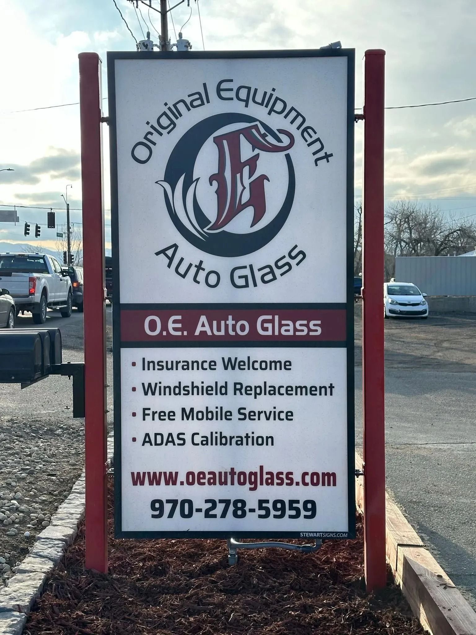 A rectangular sign for O.E. Auto Glass with contact information, services, and logo, set against a gravel roadside.