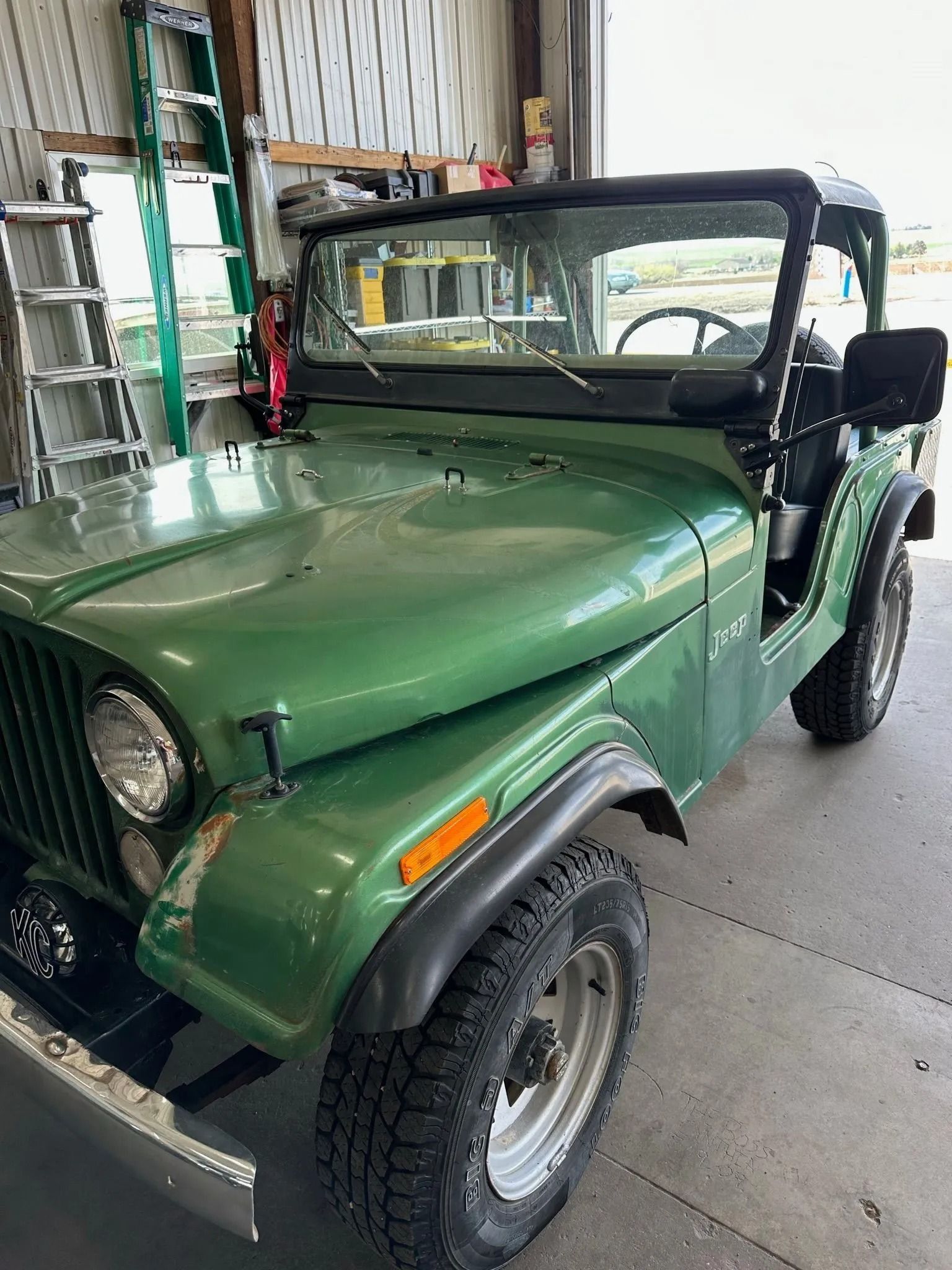 A green vintage Jeep CJ parked in a garage with a ladder visible in the background.