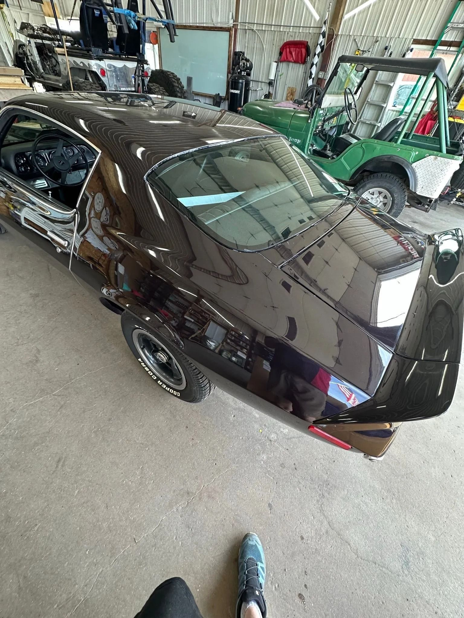 A shiny dark brown vintage muscle car parked indoors, with a green utility vehicle visible in the background.