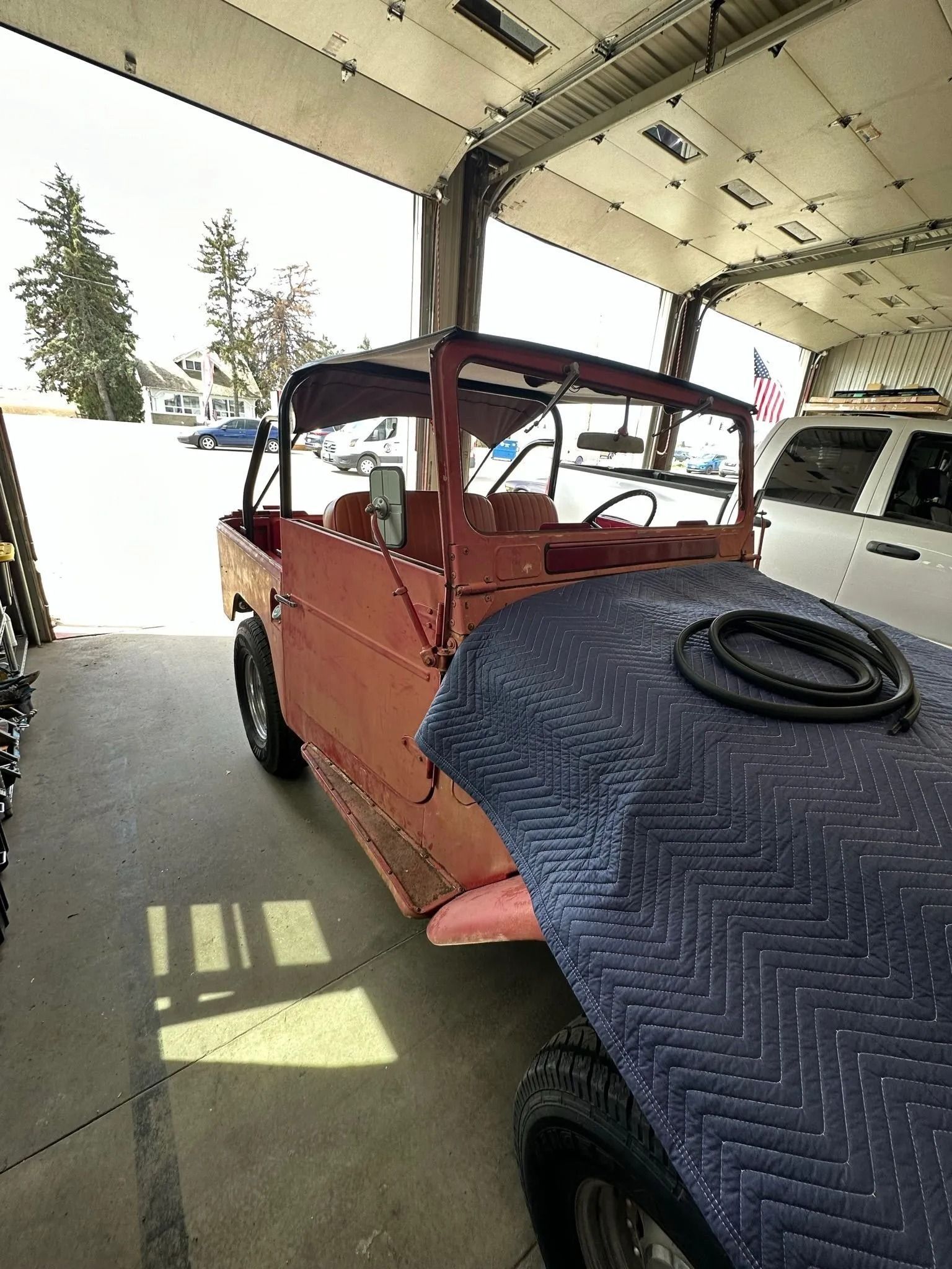 A red, partially restored vintage off-road vehicle sits inside a garage, with a blue moving blanket covering the hood.