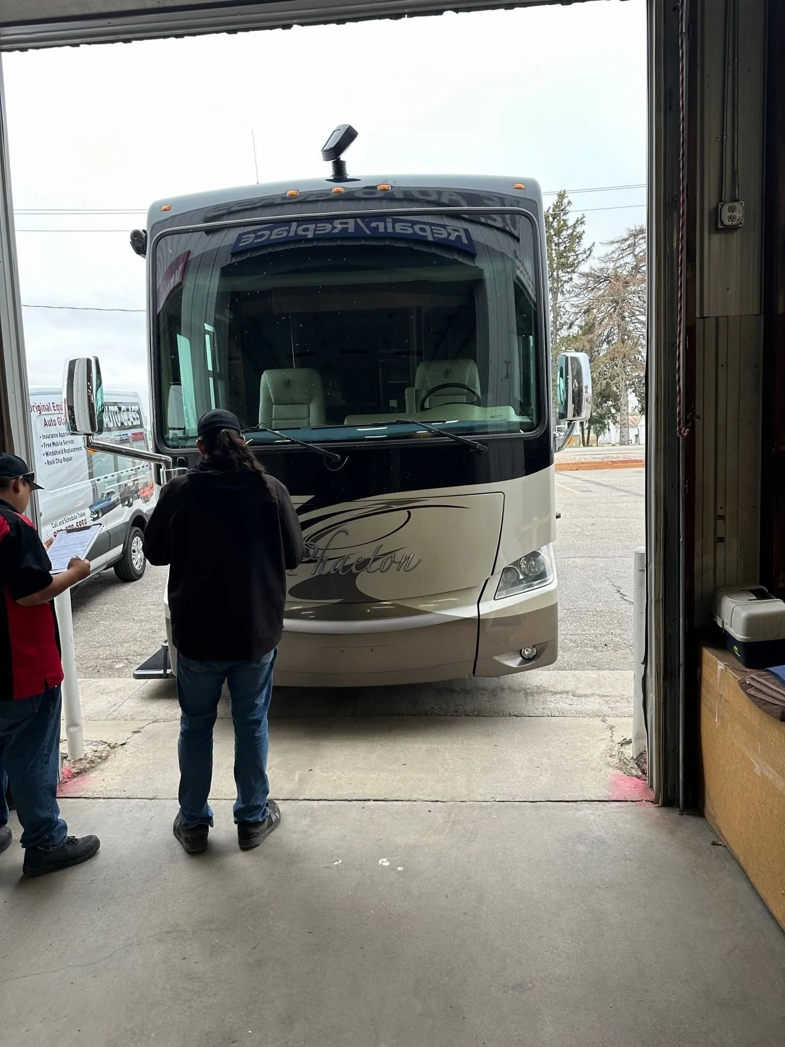 Two people stand in an open garage doorway looking at a large, tan and black recreational vehicle parked outside.