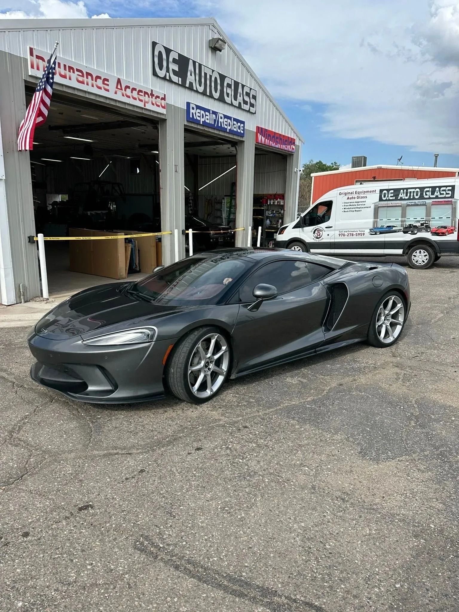 A grey McLaren GT parked outside an auto body shop with a white service van in the background.