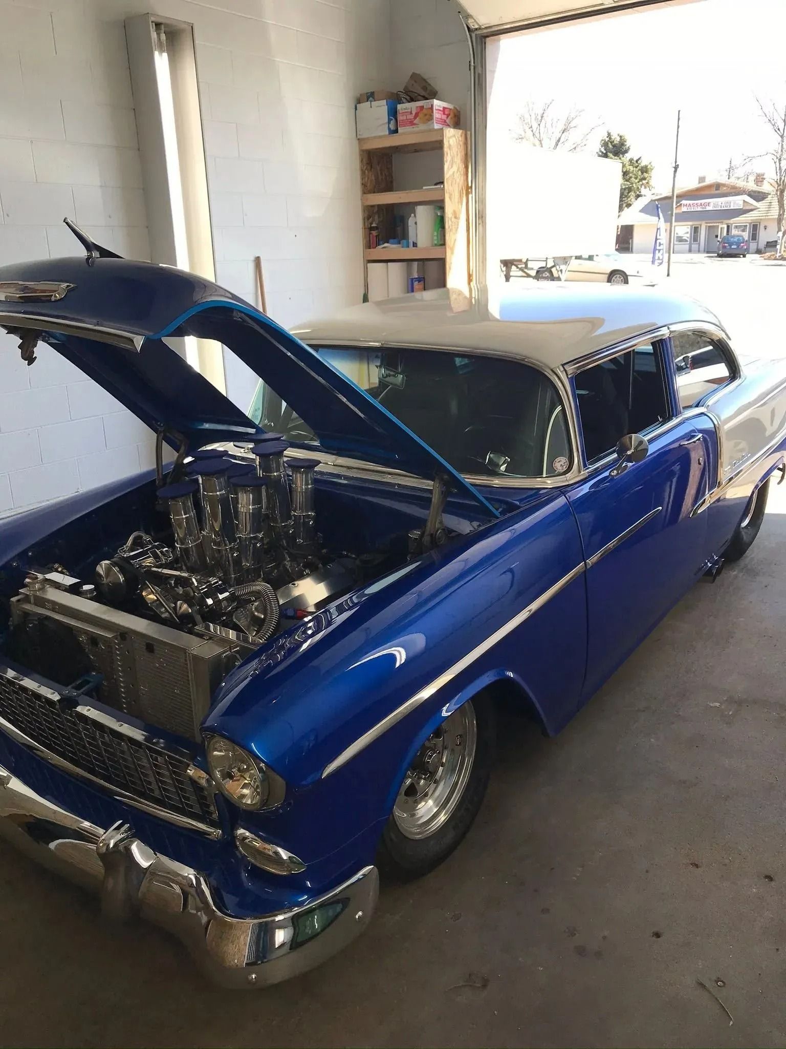 A bright blue classic 1955 Chevrolet with an open hood and a chrome engine, parked inside a garage.