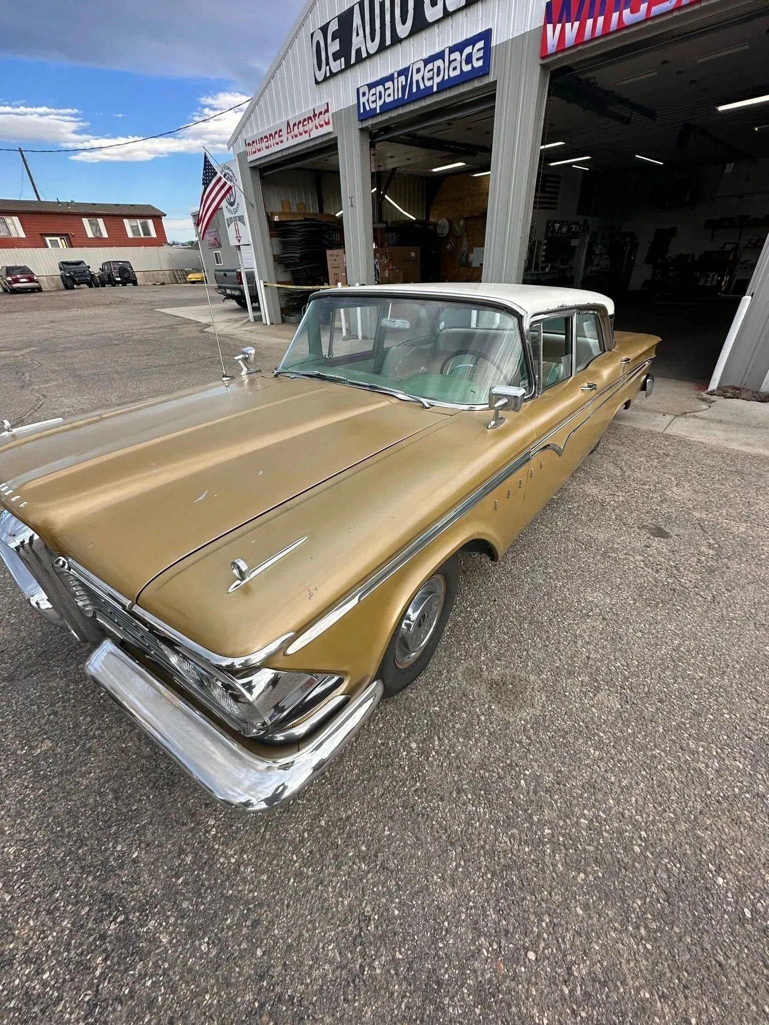A gold, vintage sedan with a white roof is parked on a gravel lot in front of an auto repair shop.