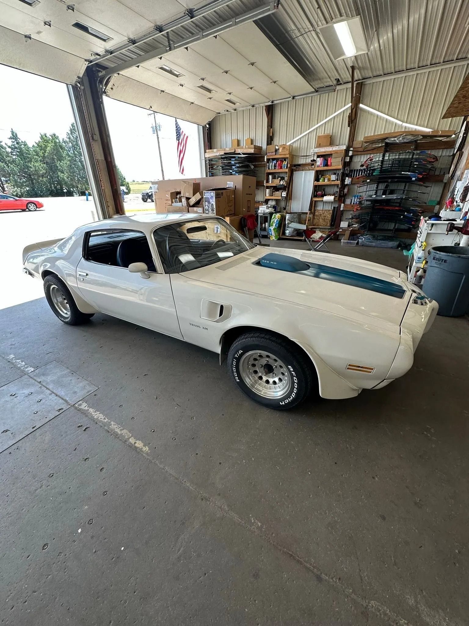 A white vintage Pontiac Firebird with a blue racing stripe parked in a garage.