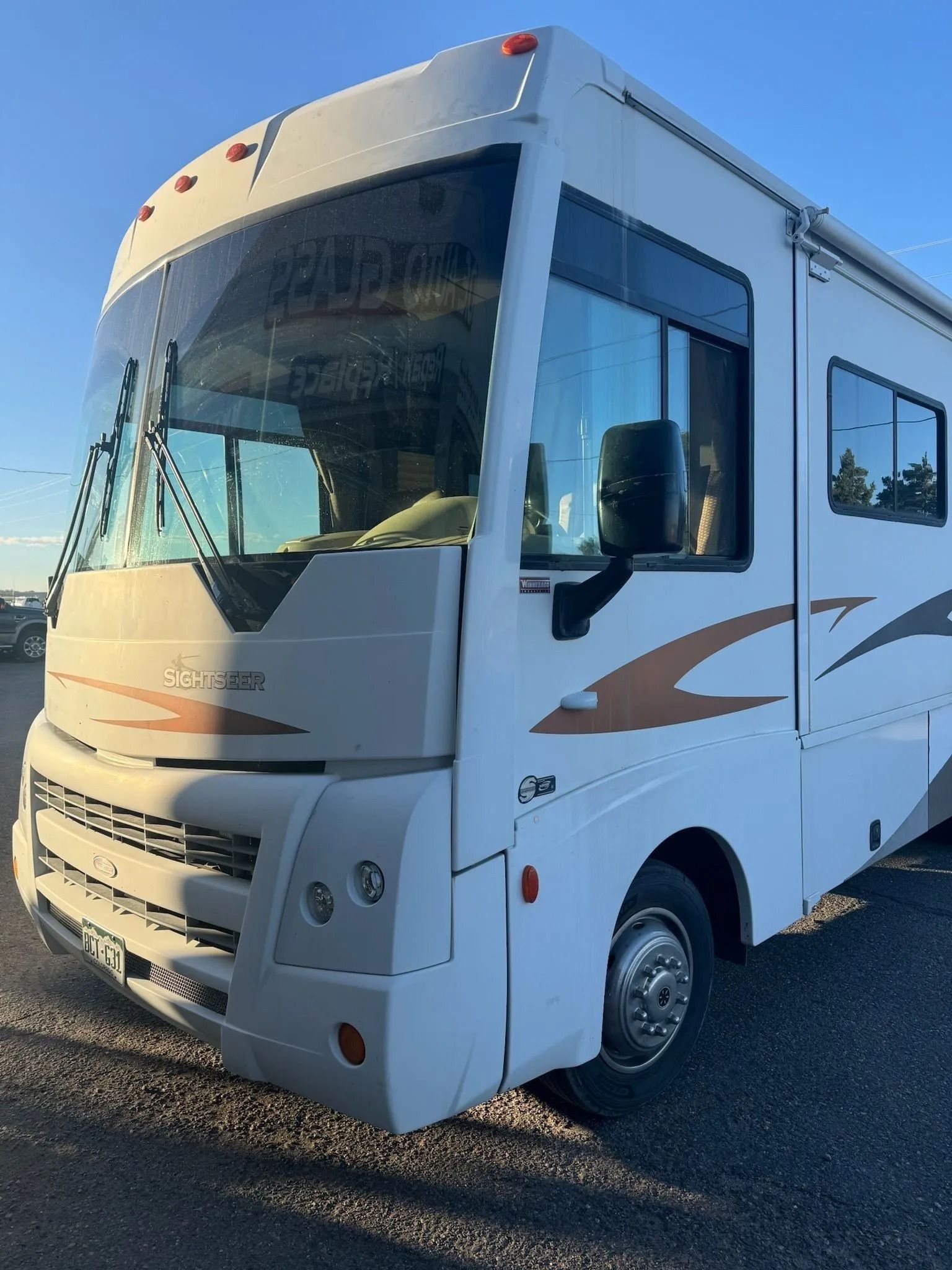 A white Class A motorhome with bronze graphics parked on a gravel lot under a clear blue sky.