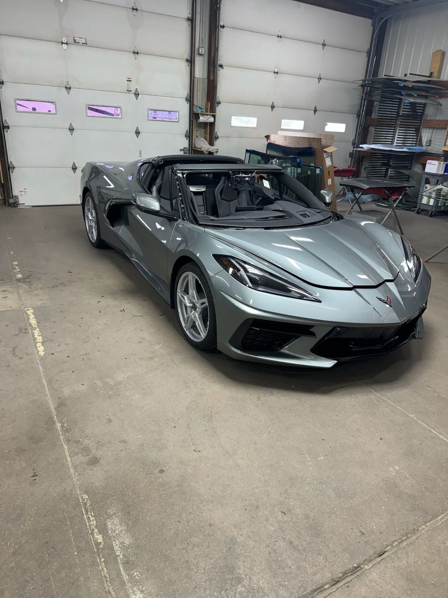 A grey, convertible Chevrolet Corvette parked inside a workshop with a large, white overhead garage door.