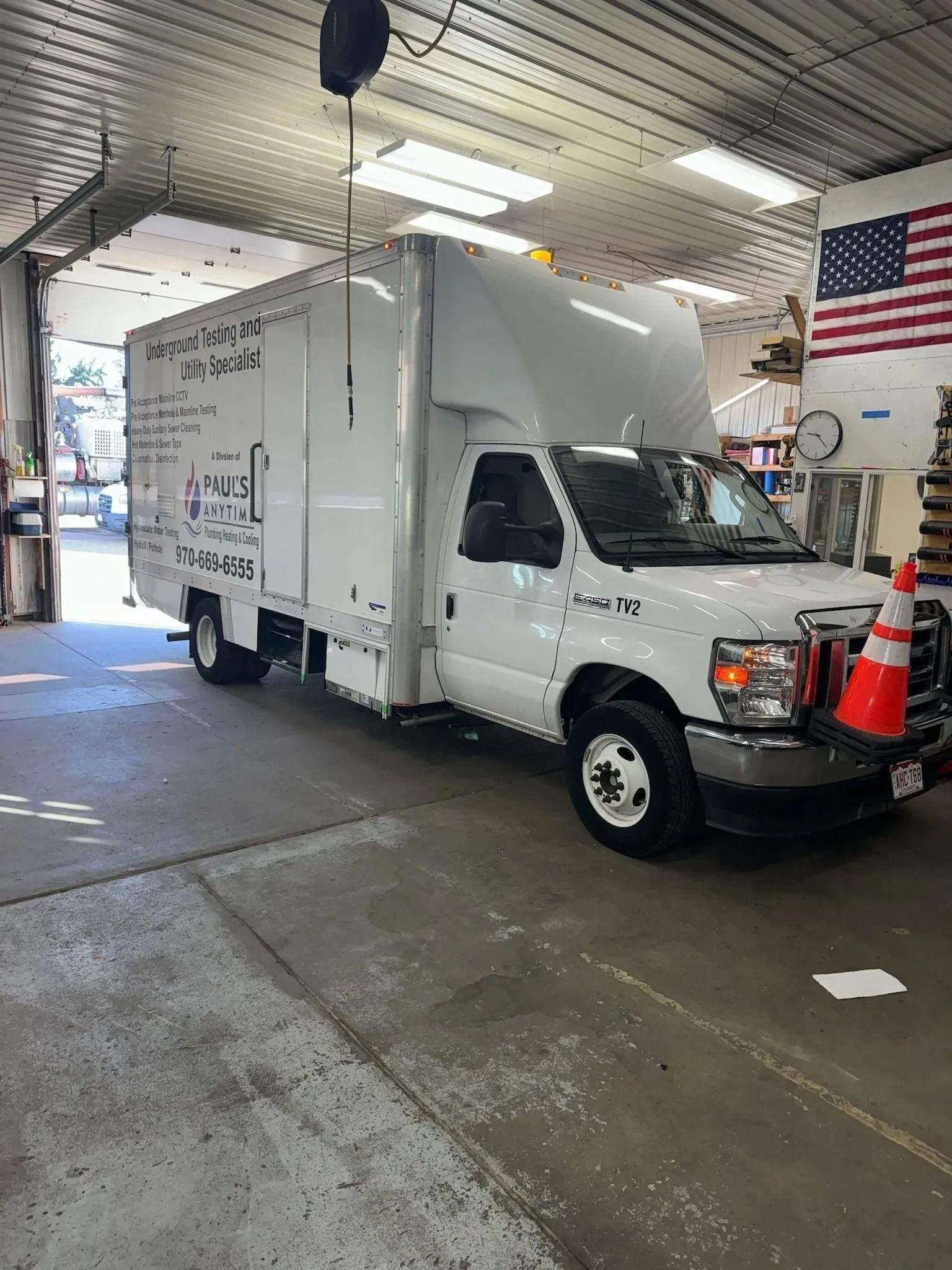 A white box truck parked inside a garage next to an American flag, with an orange traffic cone in front of the bumper.