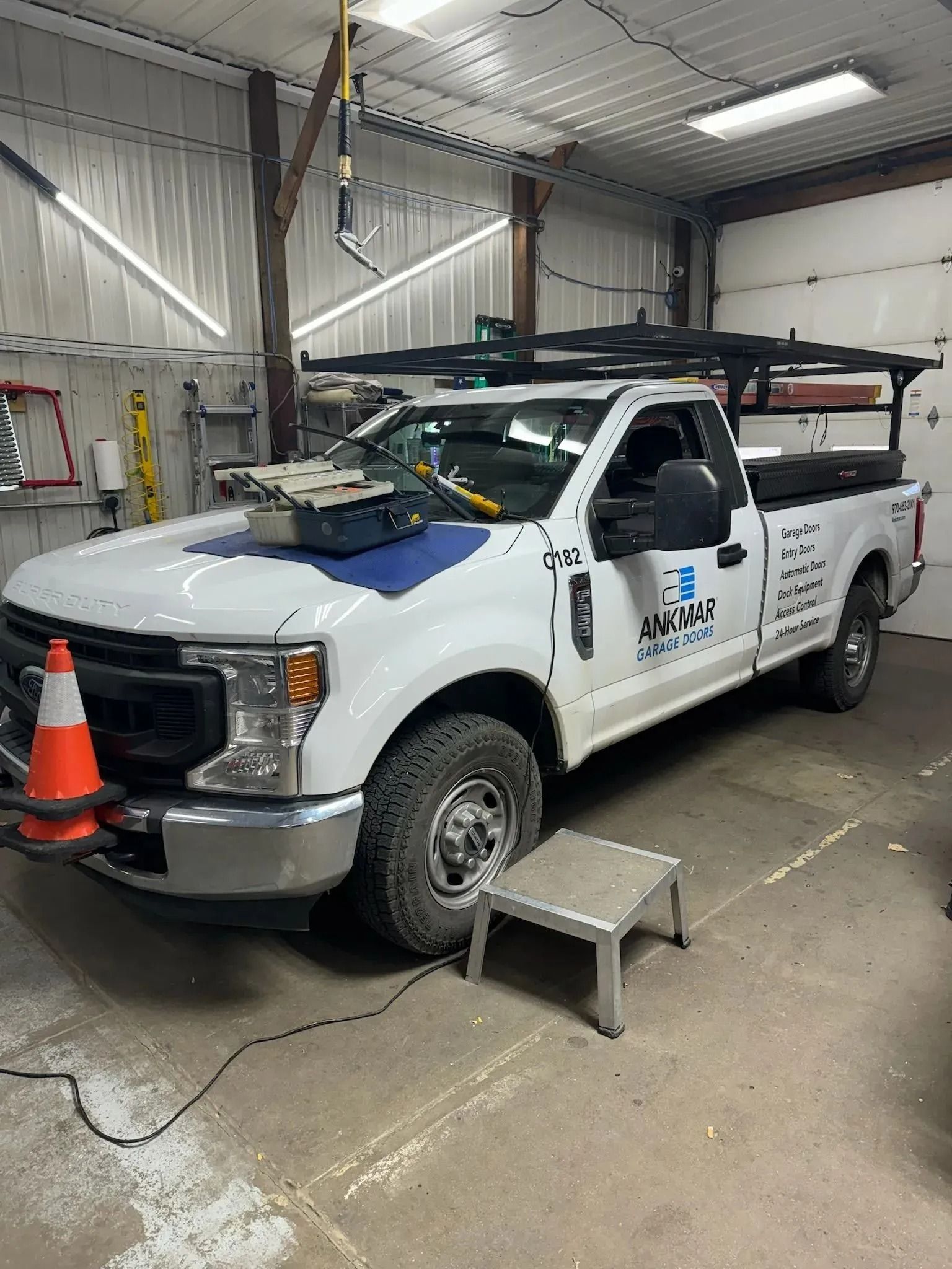 White work truck with a roof rack and company logo parked inside a garage, with an orange cone near the front bumper.