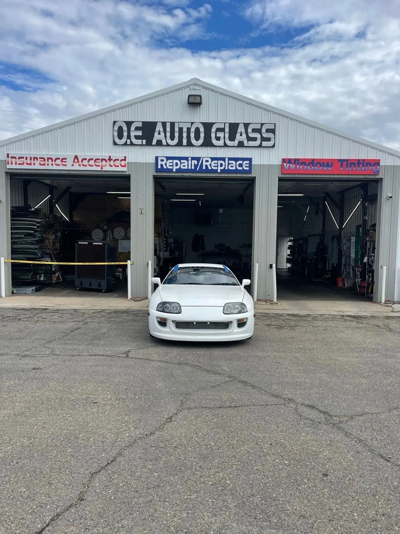 A white sports car parked in front of Q.E. Auto Glass, a repair shop with three open garage bays.