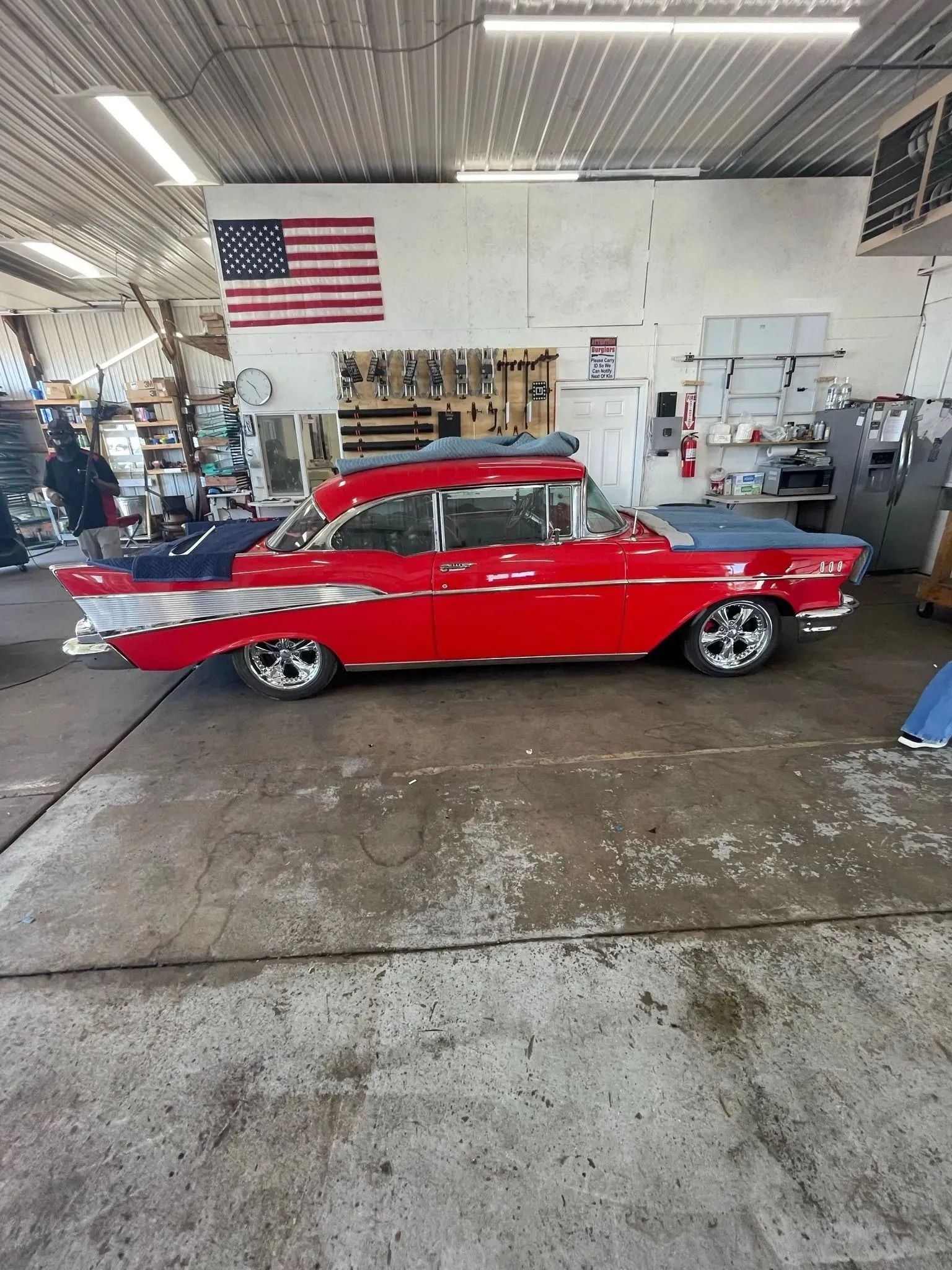 A bright red vintage car with chrome accents parked inside a garage, with an American flag hanging on the back wall.