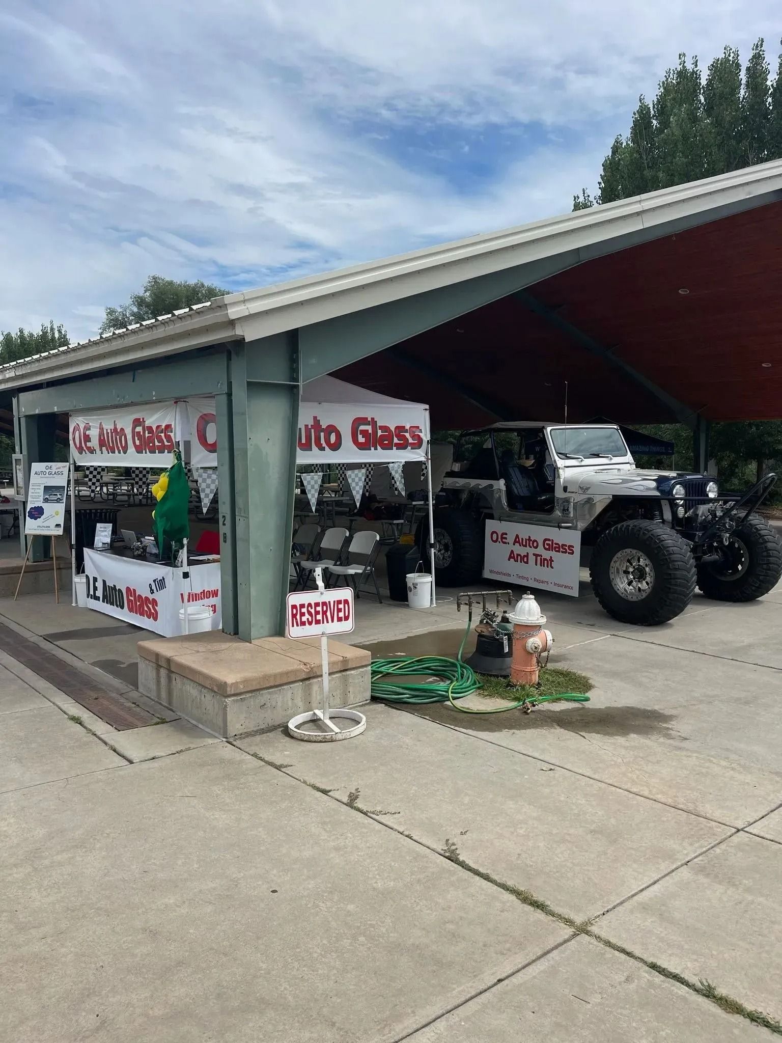 A mobile auto glass repair stand set up under a covered pavilion next to a large, white off-road vehicle with big tires.