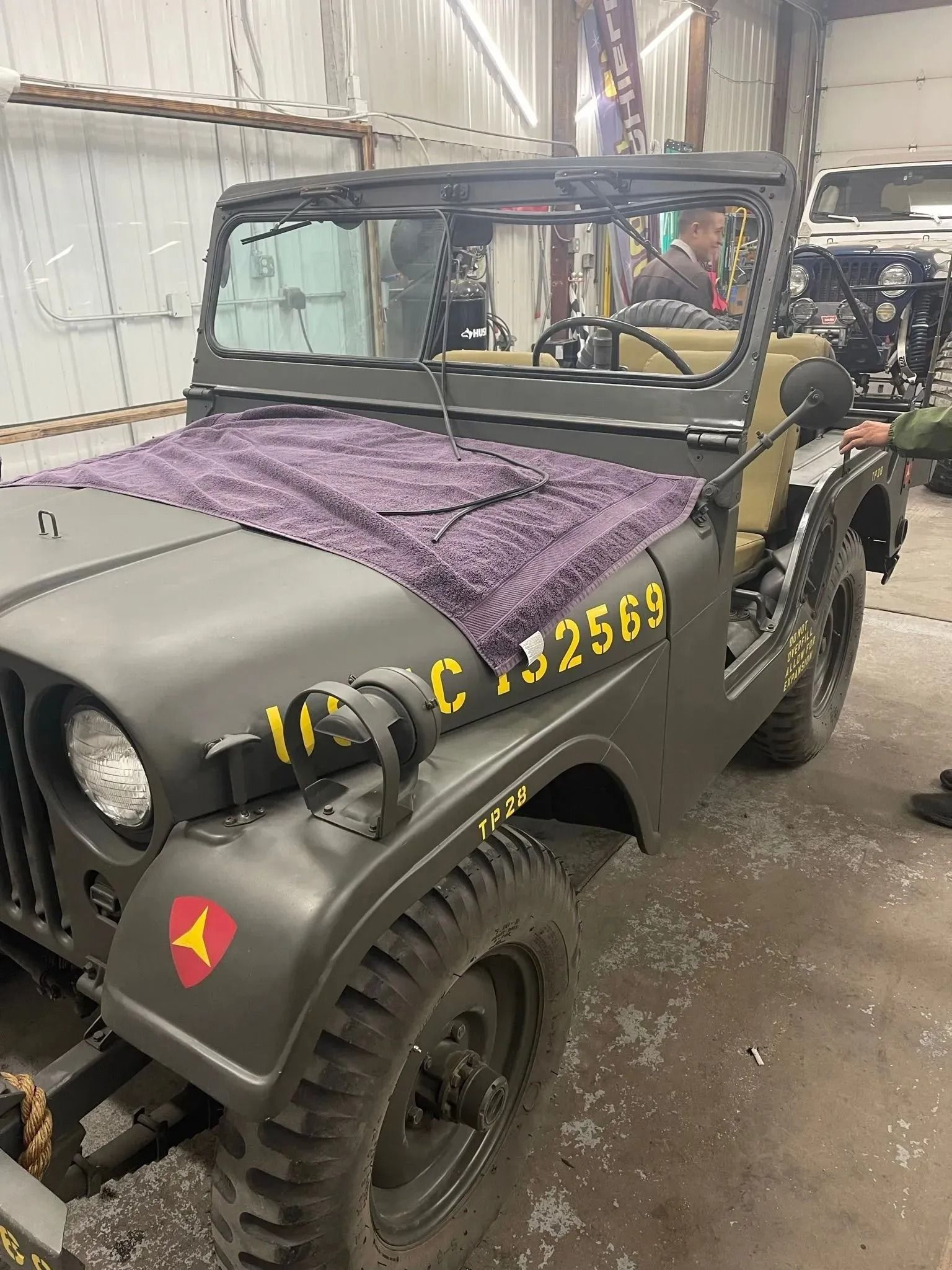 A restored olive drab military Jeep with yellow markings, parked indoors in a workshop setting.