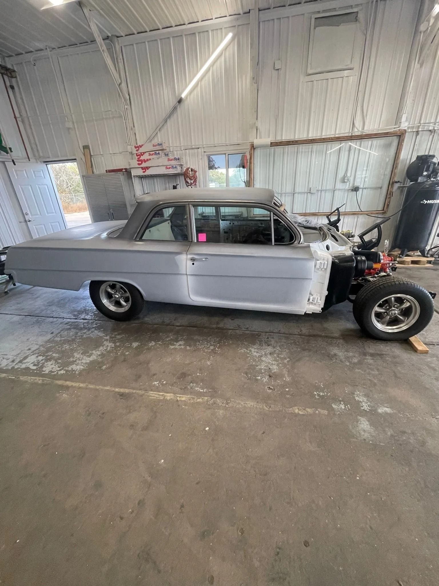 A grey, stripped-down vintage coupe car parked in a workshop garage, missing its front fenders and hood.