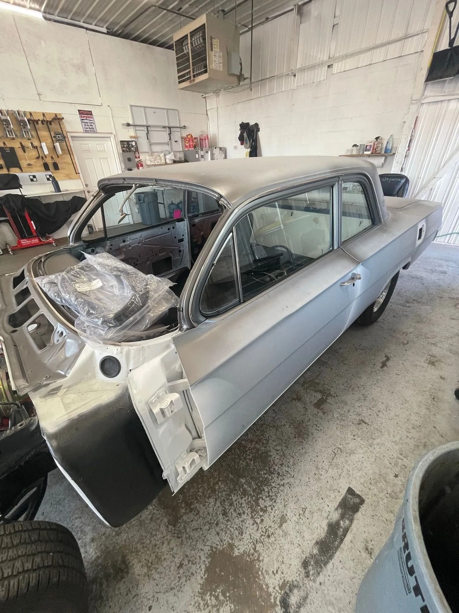 A grey classic car body under restoration inside an automotive repair shop with tools and equipment in the background.