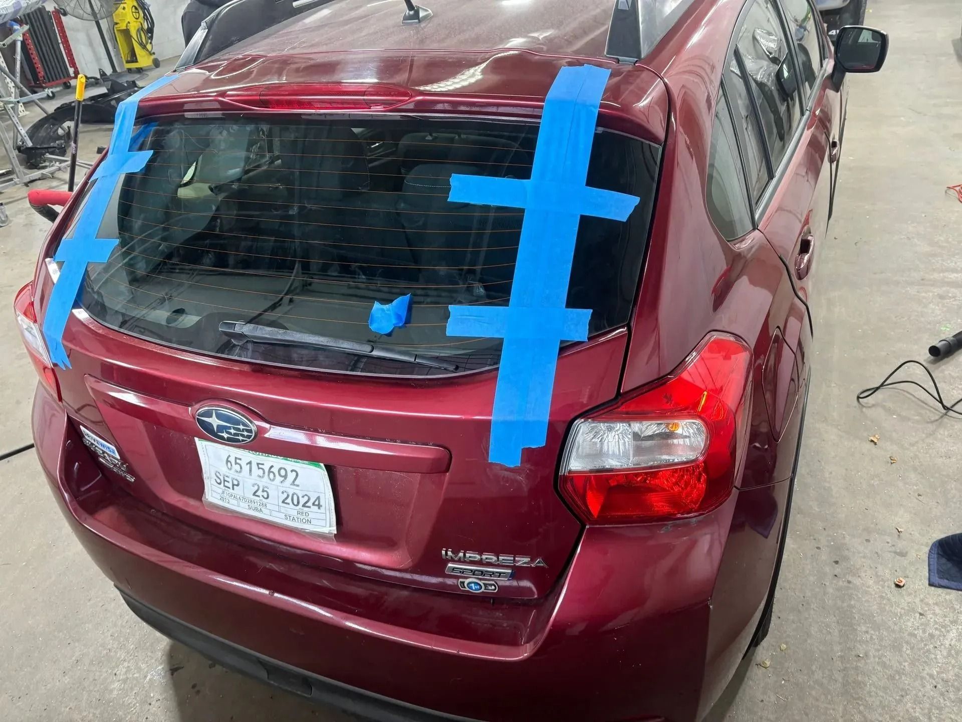 A red hatchback in a repair shop with blue tape securing the rear window glass.