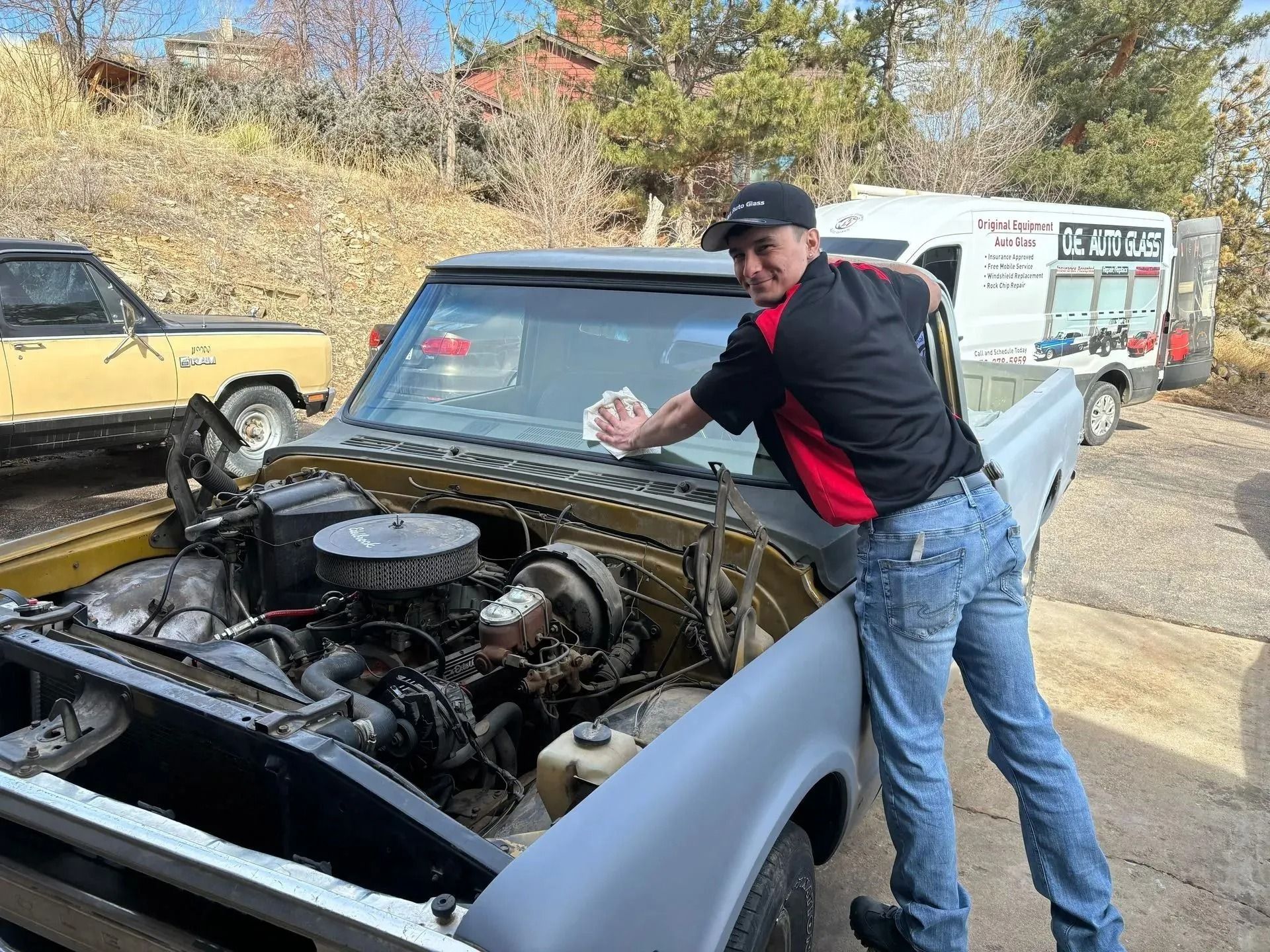 A person wearing a black and red shirt cleans the windshield of a vintage truck in a sunny, outdoor mechanic's lot.