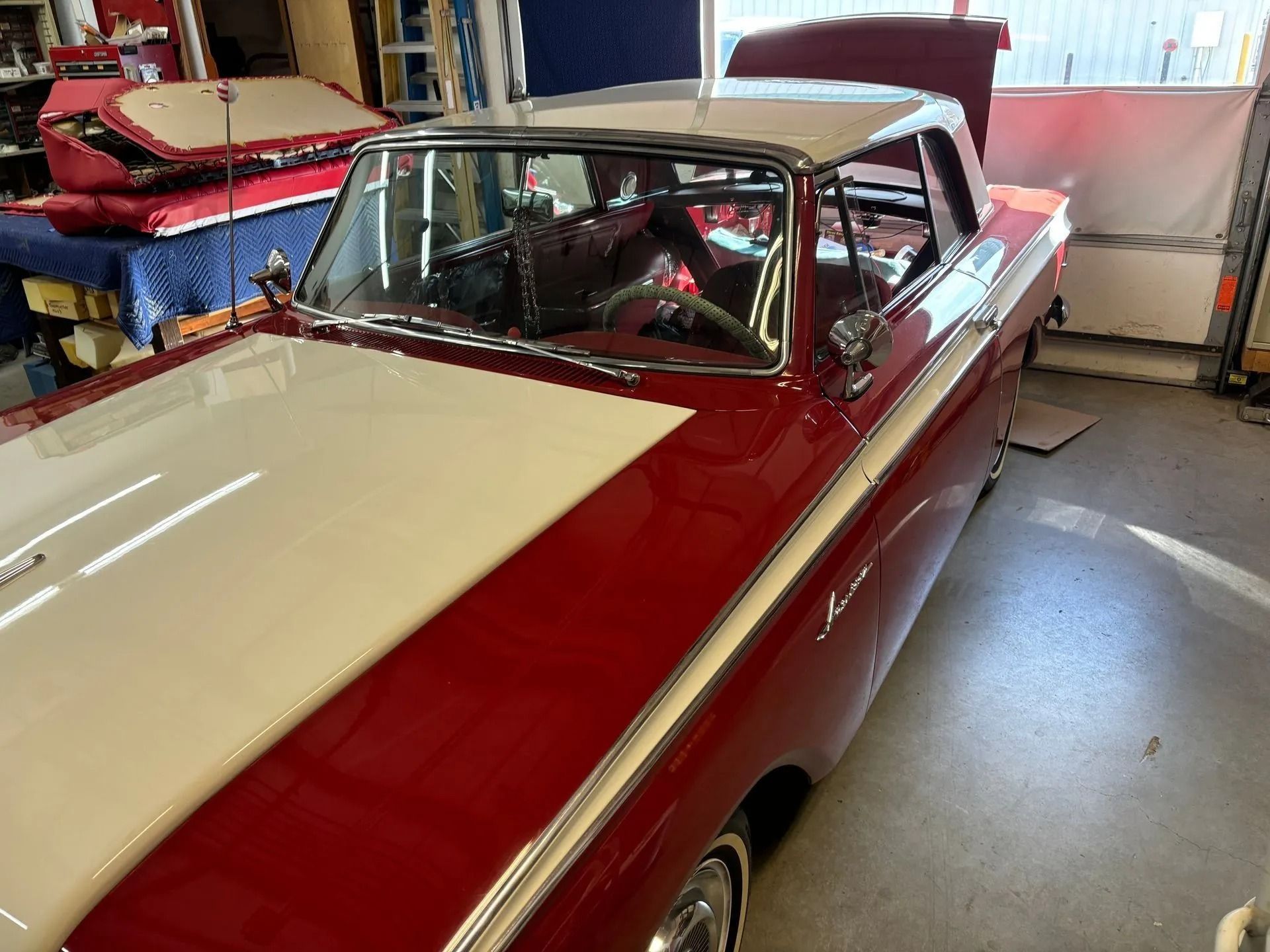 A cherry-red and cream classic car parked inside a garage with its trunk open.