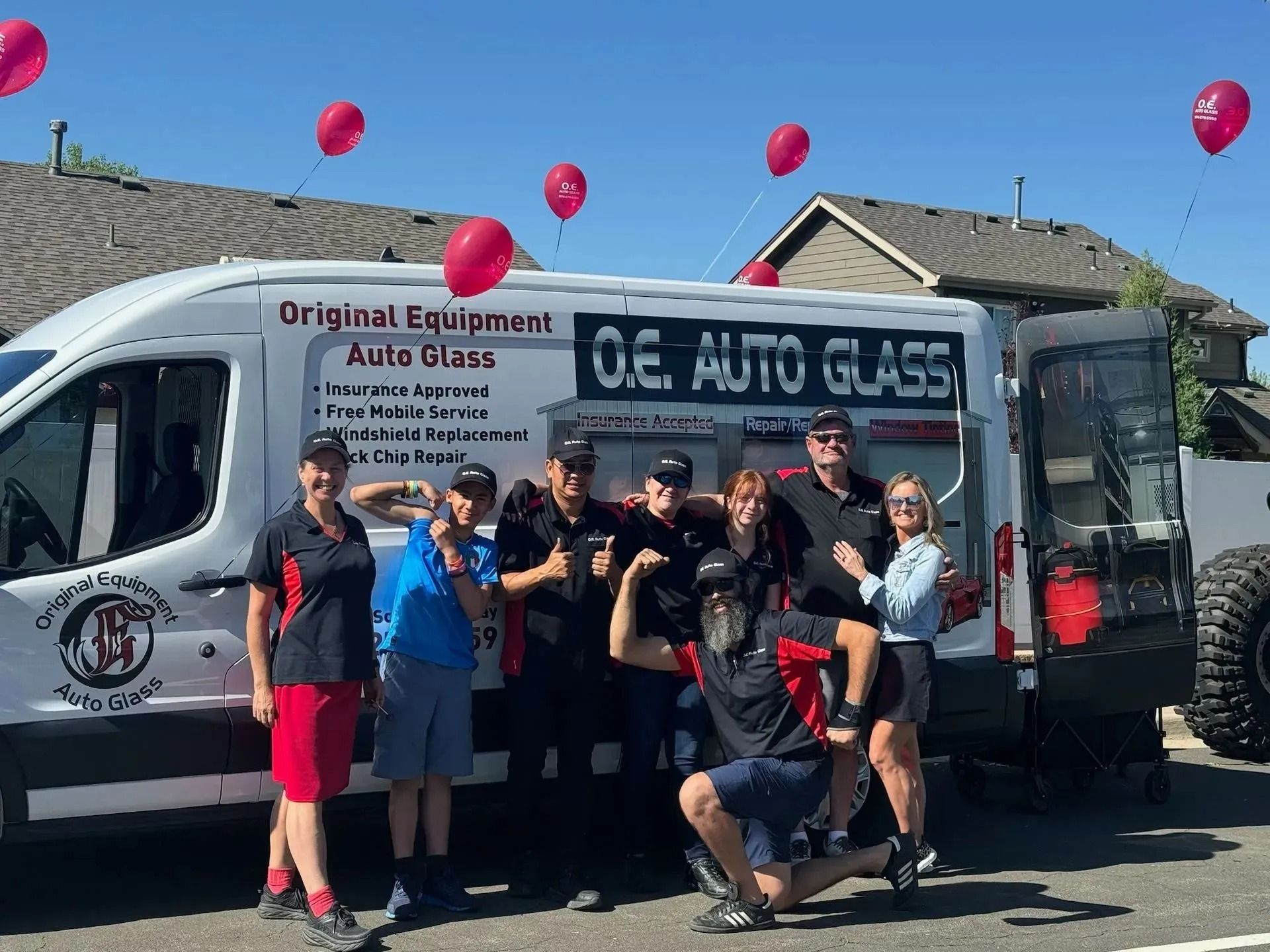 A group poses in front of an O.E. Auto Glass van decorated with pink balloons on a sunny day.