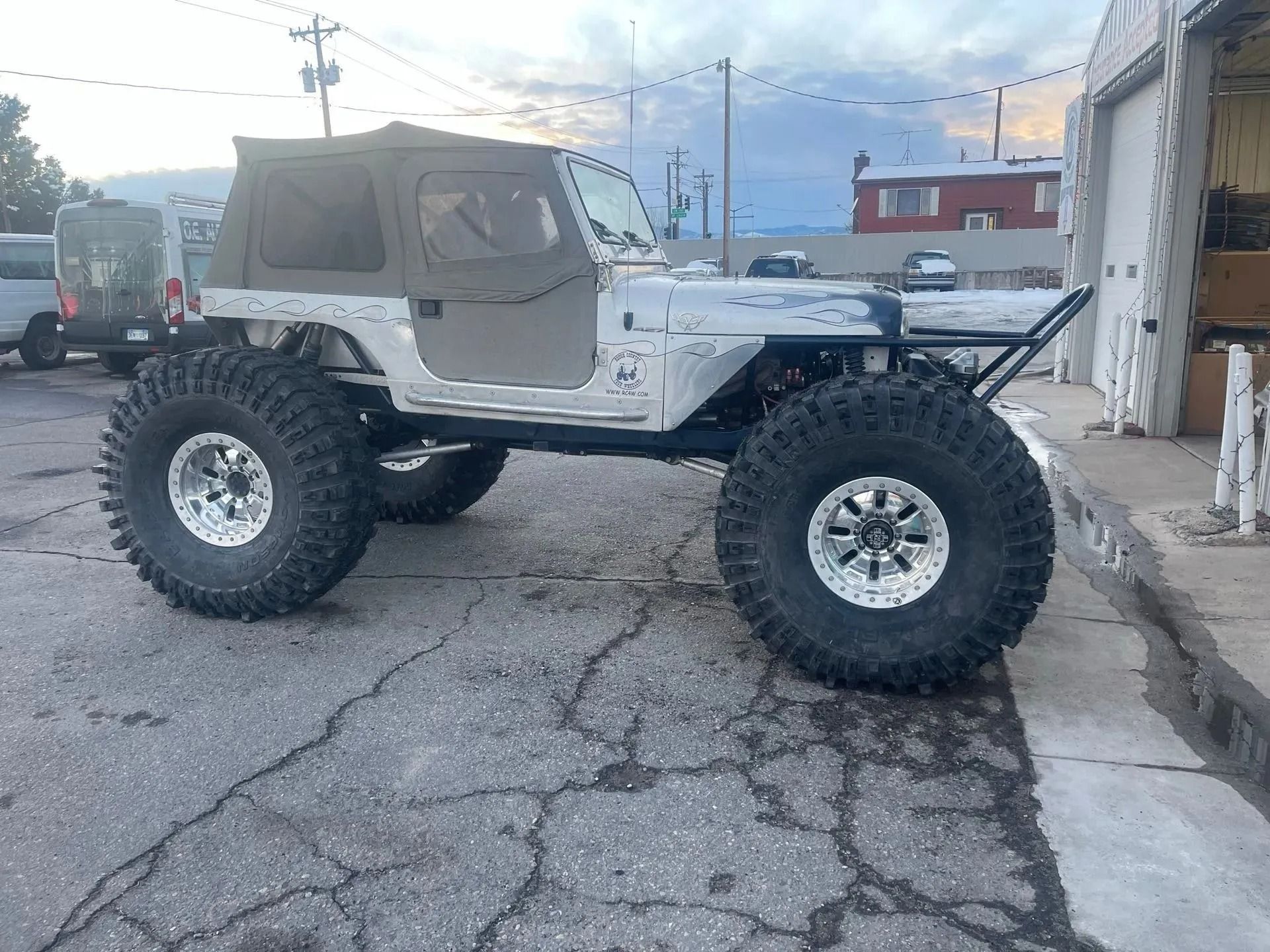 A white modified off-road Jeep with large oversized tires parked on an asphalt lot near a building.