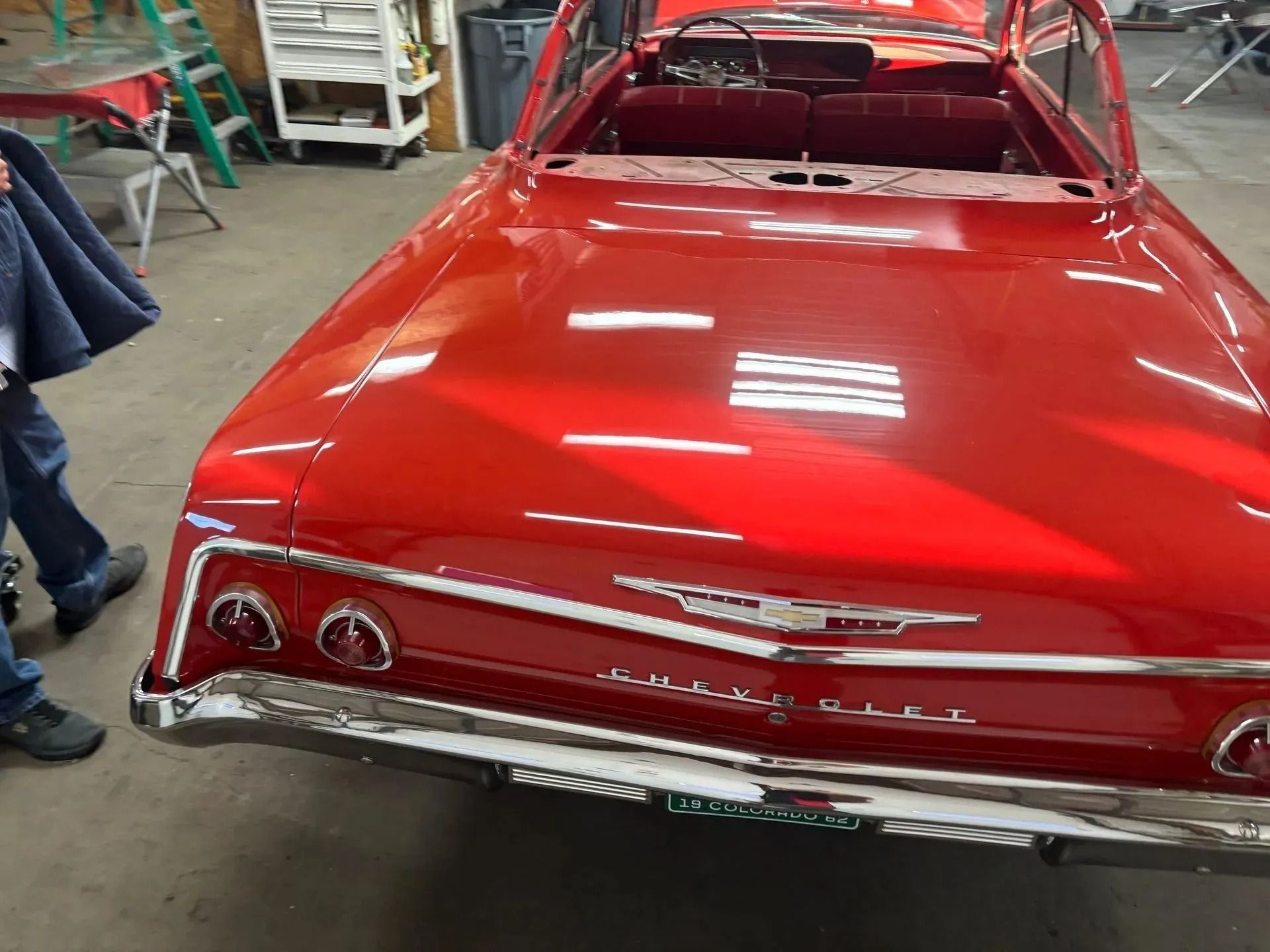 A bright red classic Chevrolet Impala in a workshop, showcasing its polished rear trunk, chrome bumper, and taillights.