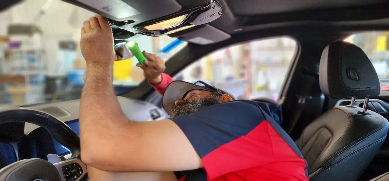 A person in a red and blue shirt uses a flashlight to inspect the interior roof area near the rearview mirror of a car.