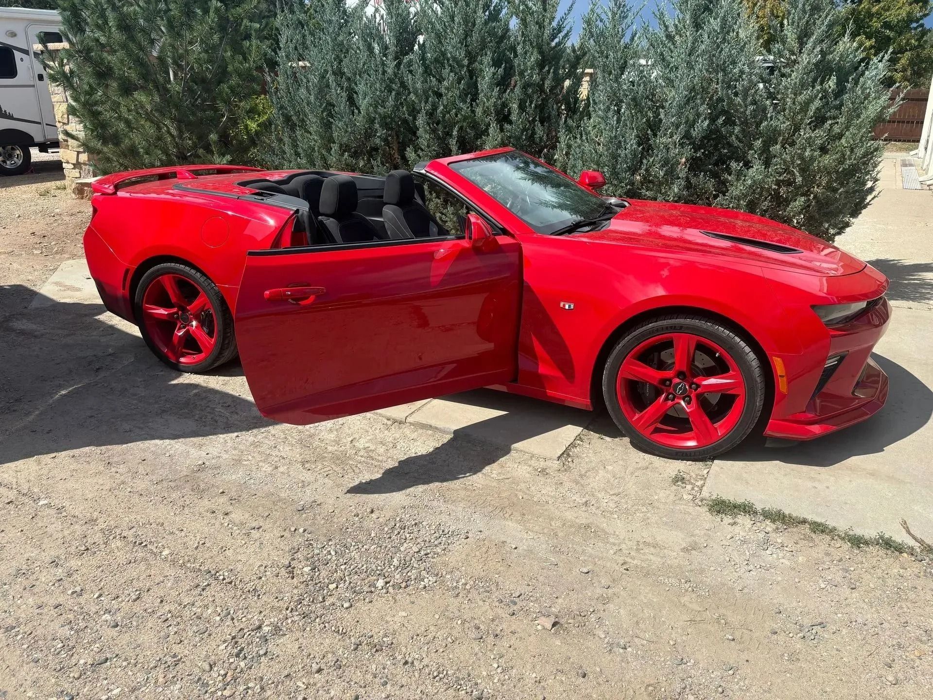 A bright red convertible Chevrolet Camaro parked on a gravel surface outdoors with its driver-side door open.