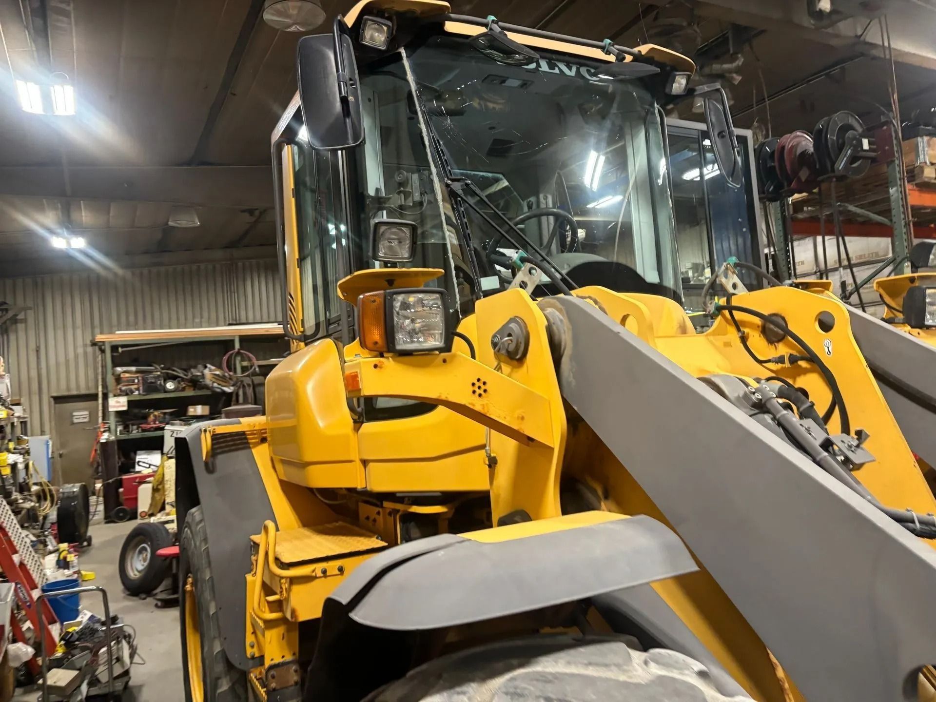 A yellow Volvo construction loader parked inside a dimly lit workshop, showing the cabin and heavy front lift arms.