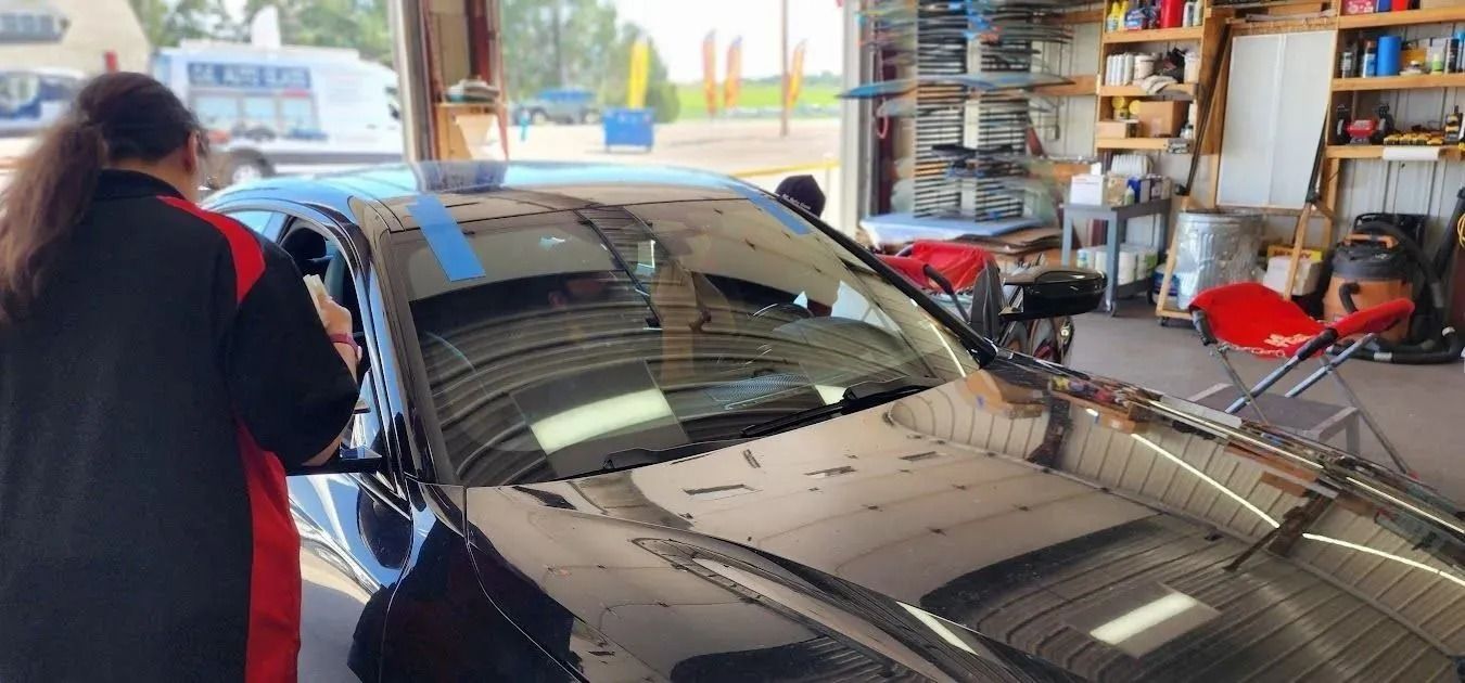 A person in a black and red uniform works on the windshield of a black car inside an automotive service garage.