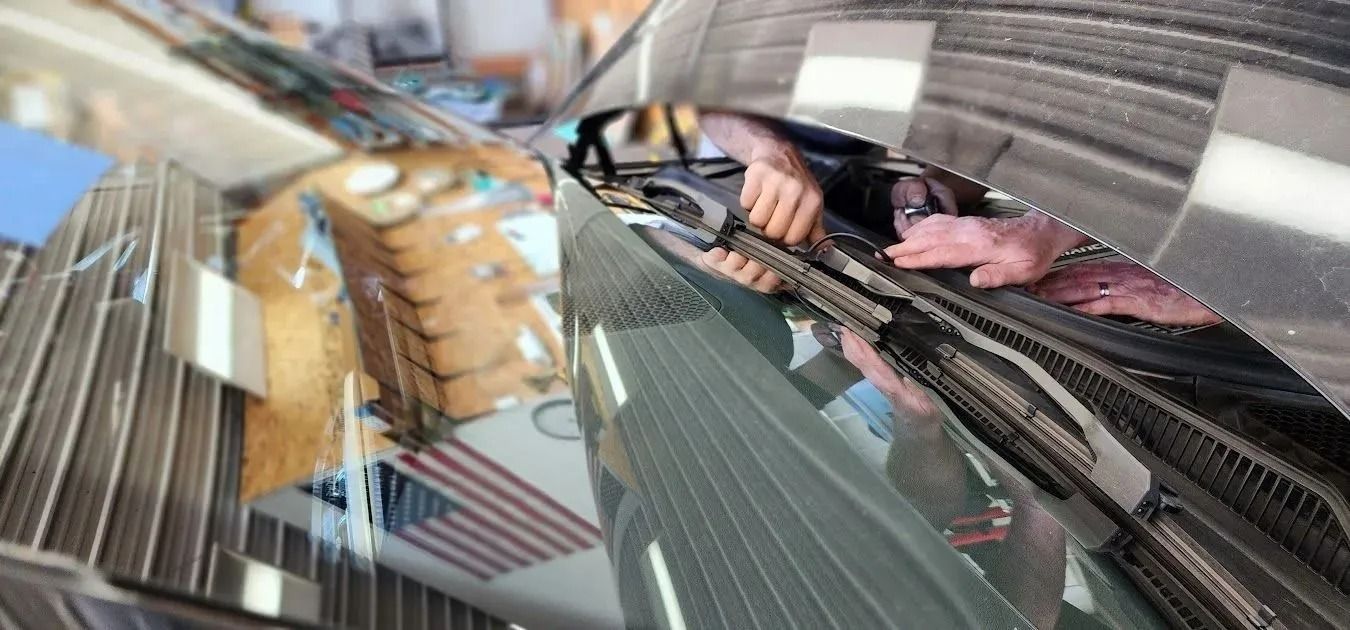 Hands work on the internal mechanisms under the hood of a car, seen through a clean windshield with a reflection of a flag.