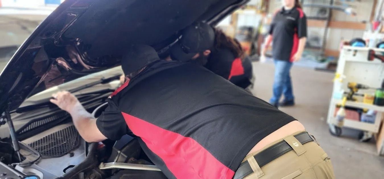 Two people in black and red uniforms work on an open car hood in a workshop, while another person stands nearby.