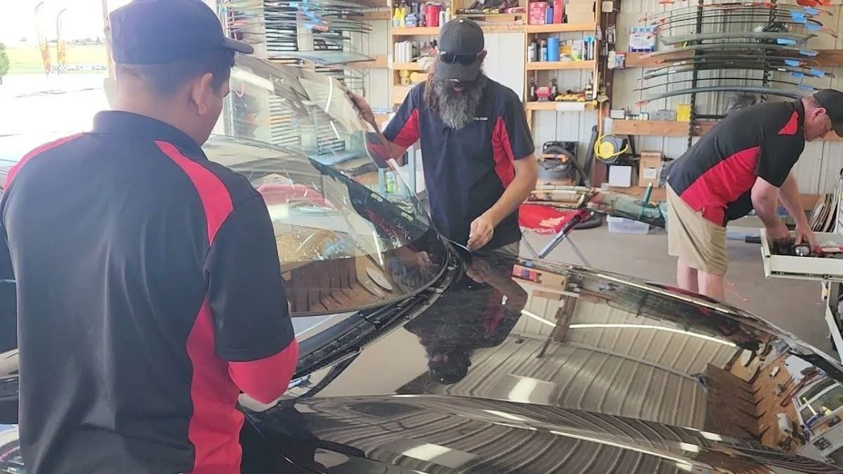 Three technicians in red and black uniforms work together to install a windshield on a car inside an auto shop.