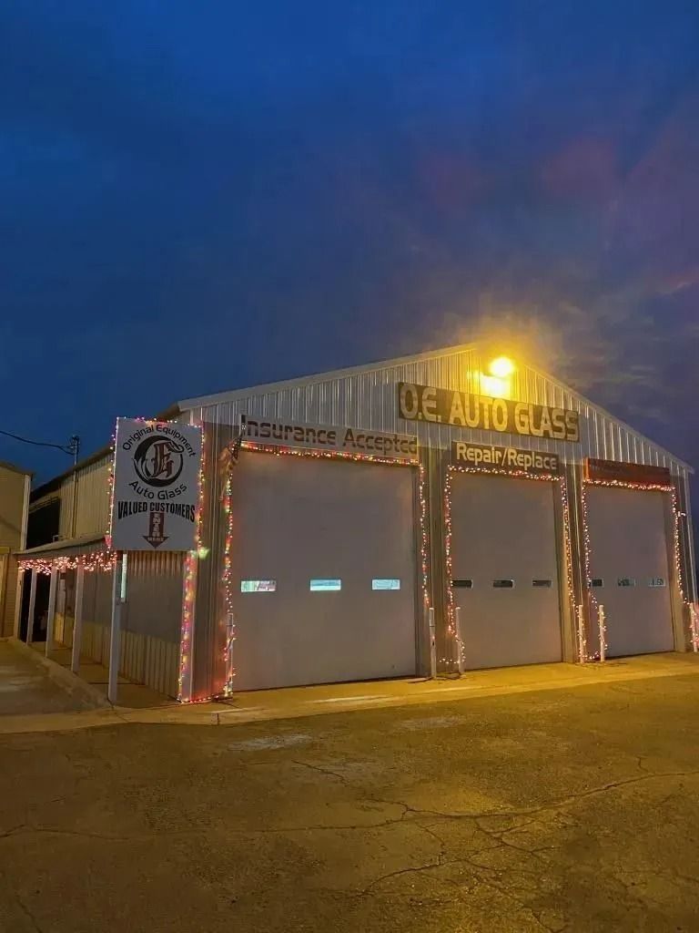 A building labeled OE Auto Glass at dusk, illuminated by exterior lights and decorative holiday string lights.