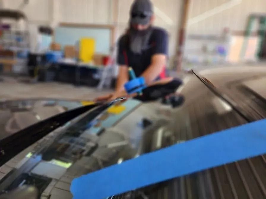 A person in a workshop uses a blue tool to work on the windshield of a car marked with blue painter's tape.
