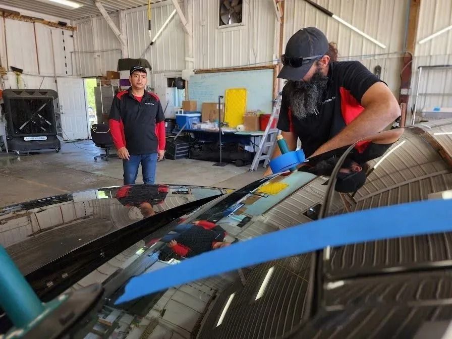 A technician works on a car windshield in a garage, while a person stands nearby watching.