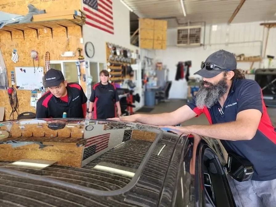 Three people in a garage workshop inspect the roof of a black vehicle; one person leans over the car while two watch.