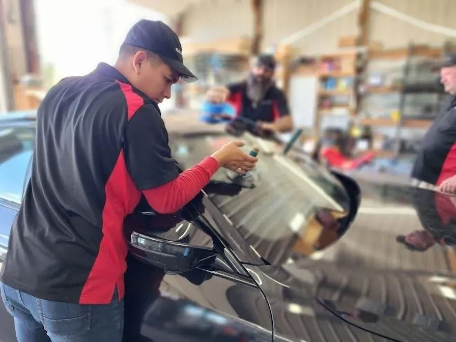 A technician in a black and red polo shirt uses a tool to repair a crack on a black car’s windshield in a garage.