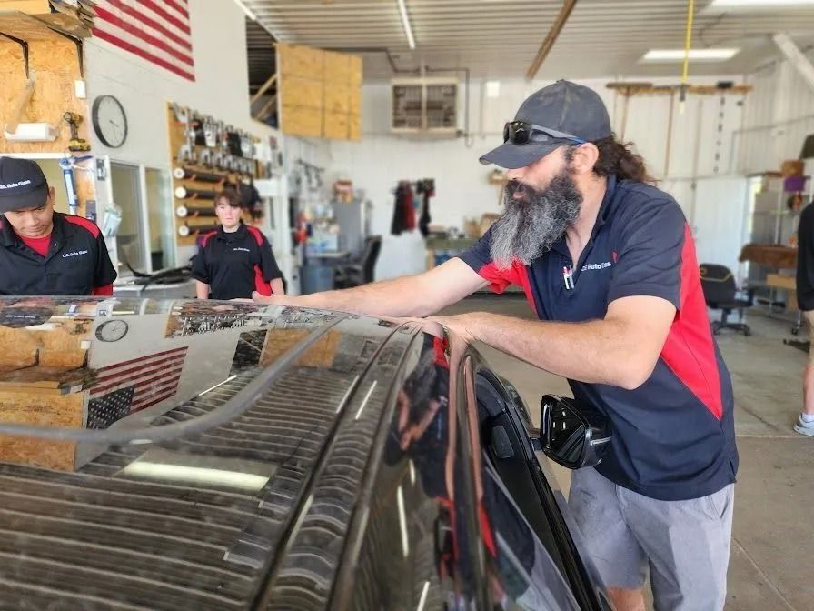 A technician with a beard and baseball cap inspects the roof of a black car in a well-lit auto repair shop.
