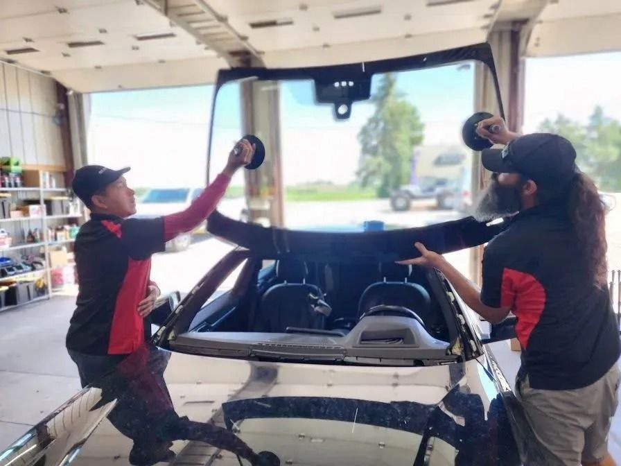 Two technicians in matching red and black uniforms lift a large replacement windshield onto a car in a repair shop.