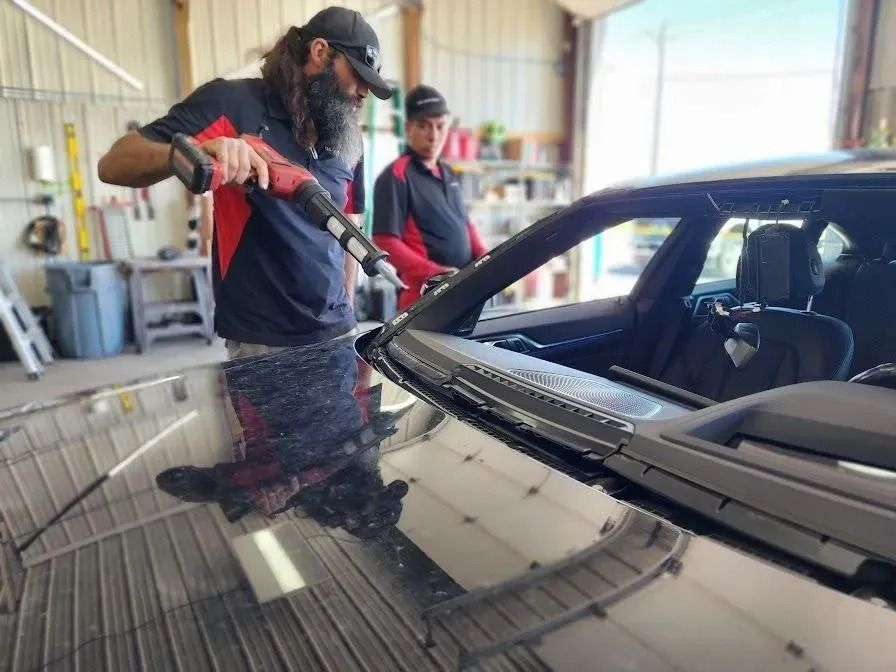 Two technicians in a garage using a specialized adhesive gun to apply sealant to a car's windshield frame.
