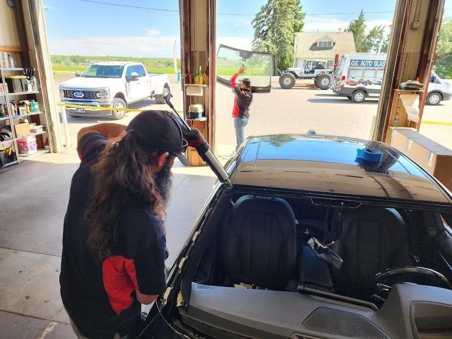Two technicians work on installing a new windshield in an automotive shop, with vehicles visible outside the garage bay.