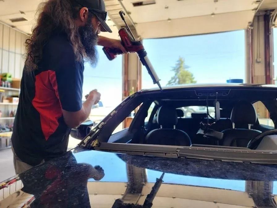 A person with a long beard uses a power caulk gun to apply adhesive to the windshield frame of a car in a garage.