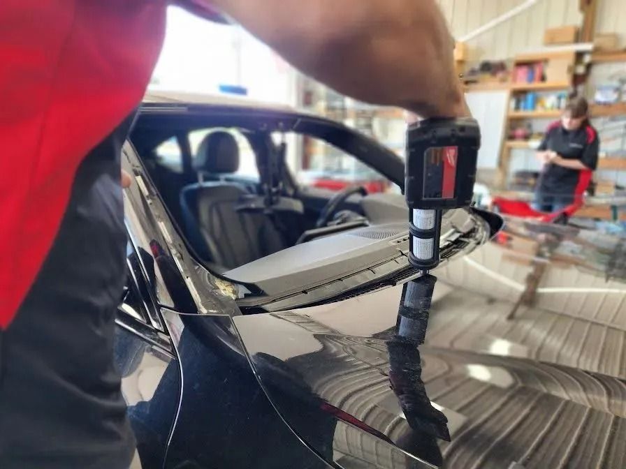 A worker in a red shirt uses a power caulking gun to apply adhesive to a car windshield frame in an auto shop.