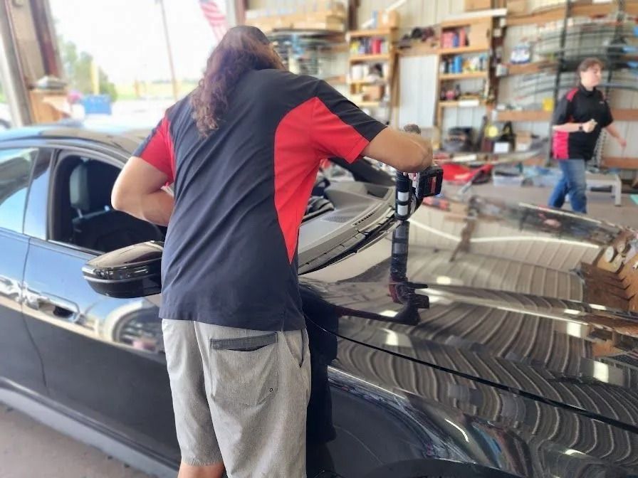 A technician uses a buffing tool on the hood of a black car in an auto shop while another person walks in the background.