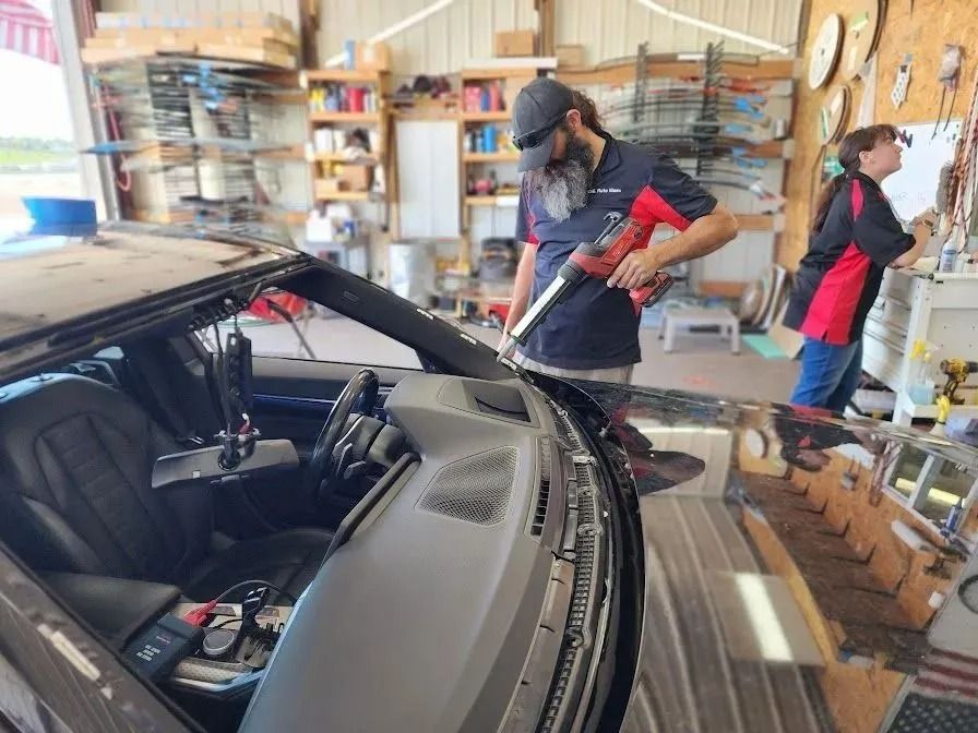 Two people working in a repair shop; one uses a power tool on a car windshield while the other looks at a whiteboard.